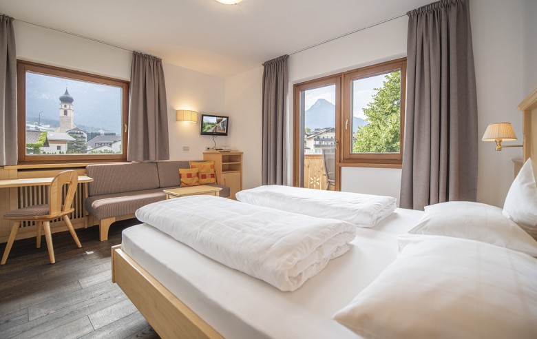 Das Zimmer mit einem Doppelbett, einer Couch, einen TV und einen Schreibtisch. Durch die Fenster sieht man die Aussicht auf das Dorf und die umliegende Natur.