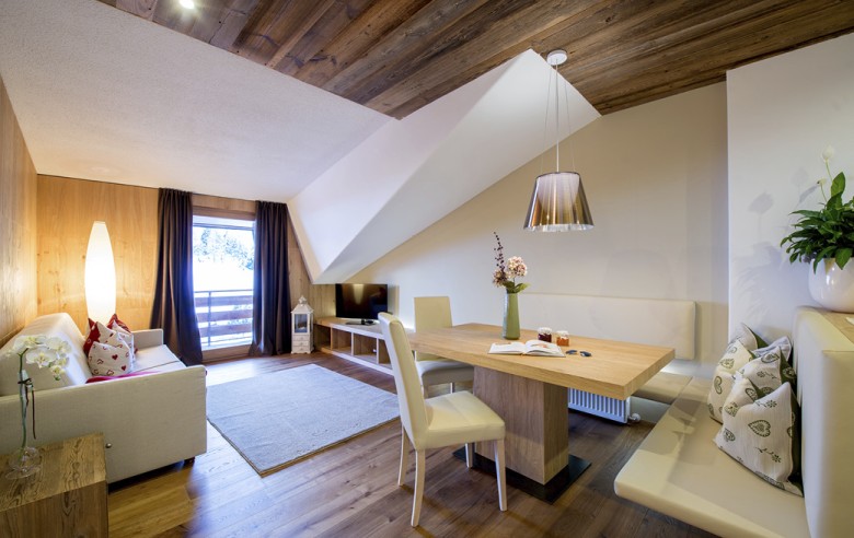 Wohnbereich der Wohnung. Man sieht einen Esstisch, einen TV und ein Sofa. Außerdem sieht man auch den Balkon und die Aussicht auf die Natur.