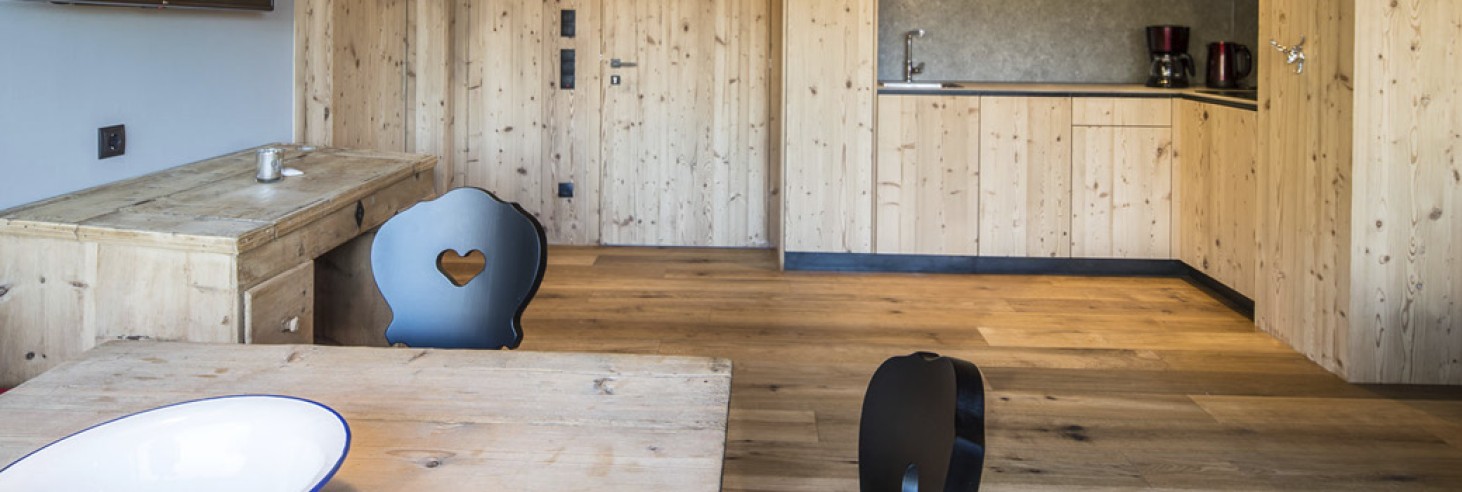 Der Wohnbereich der Ferienwohnung ist komplett in Holz gehalten und im Vordergrund sieht man den Holztisch mit schwarzen Stühlen. Im Hintergrund sieht man die Kochecke und den TV.