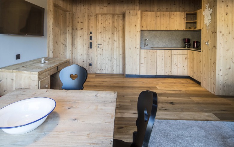 Der Wohnbereich der Ferienwohnung ist komplett in Holz gehalten und im Vordergrund sieht man den Holztisch mit schwarzen Stühlen. Im Hintergrund sieht man die Kochecke und den TV.