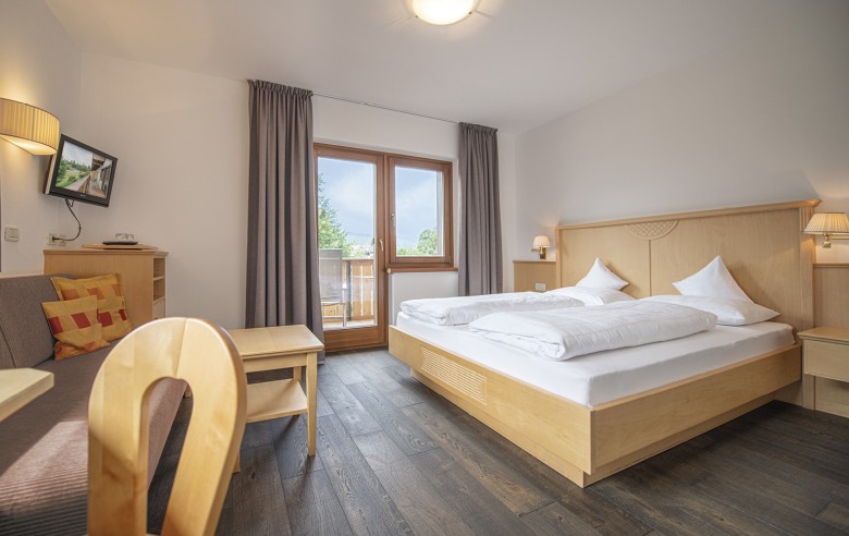 Das Komfort Zimmer mit einem Doppelbett, einer Couch und einem Fernseher. Durch die Balkontür sieht man die direkte Aussicht in die Natur.