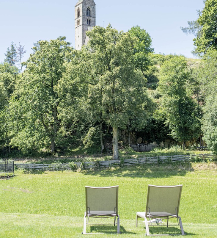 Von zwei Liegenstühlen im Garten erblickst du weite grüne Wiesen und die Kirche des Dorfes.