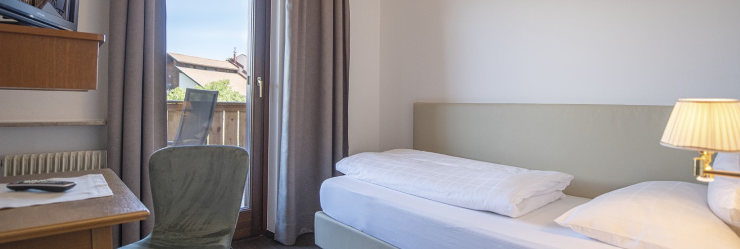 Una camera singola con letto, scrivania e televisore. È visibile anche una parte del balcone adiacente.